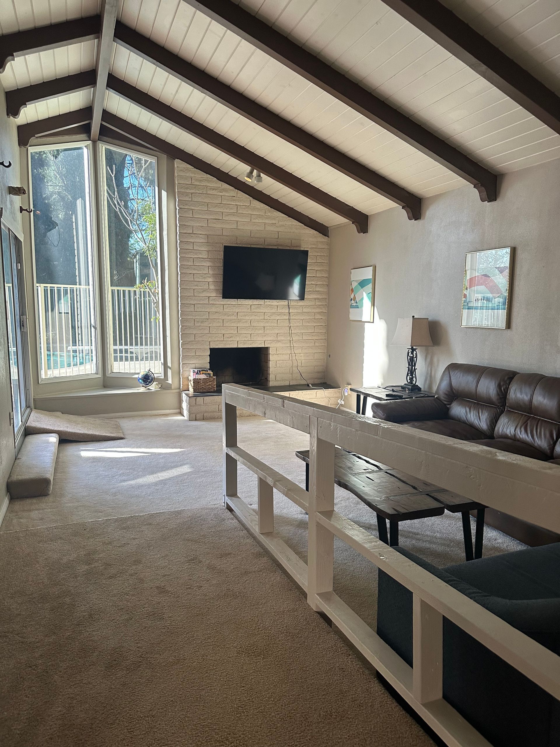 Living room with vaulted ceiling, fireplace, and large windows. Brown beams, beige carpet, dark sofa.