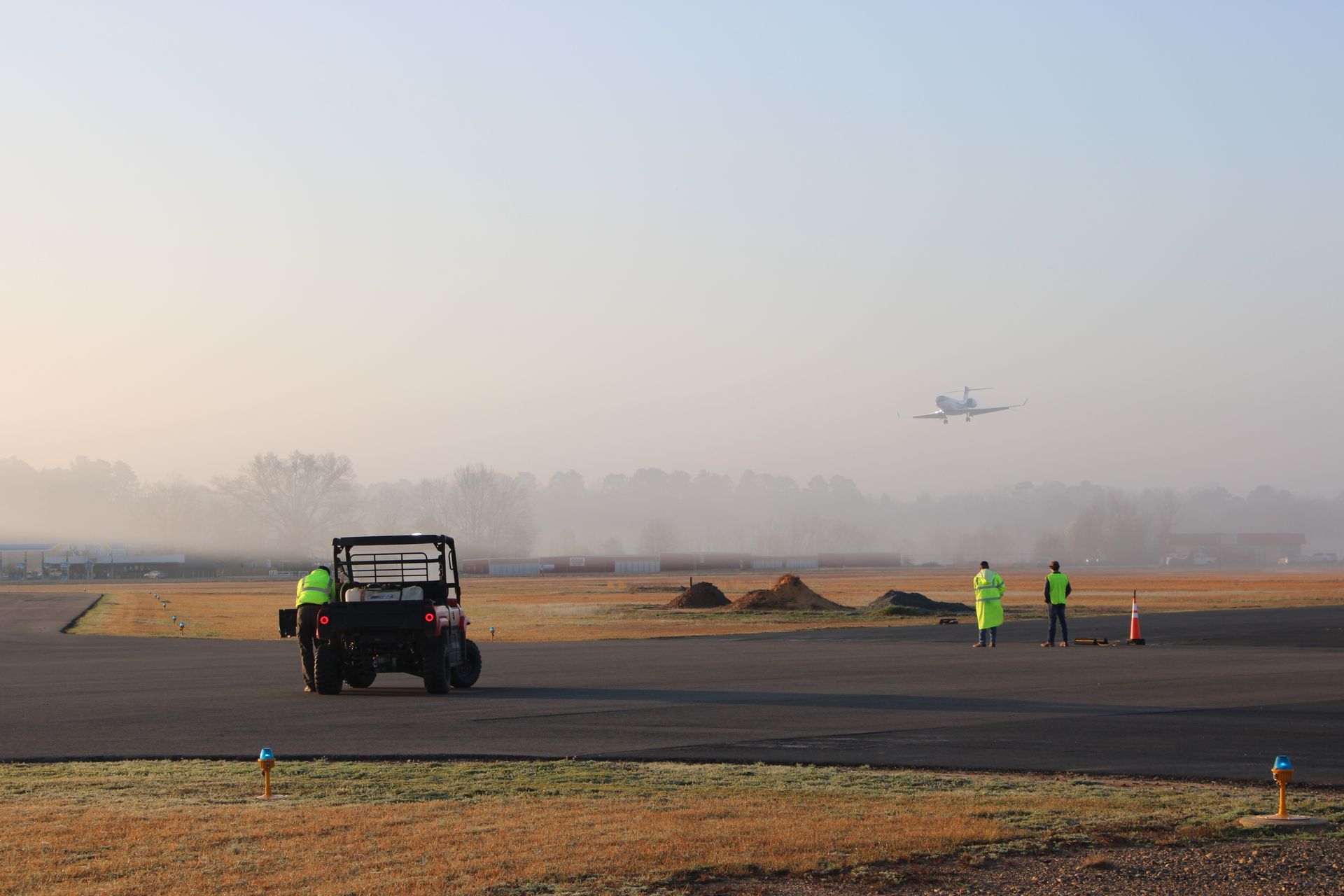 A golf cart and two workers in high-visibility vests stand on an airfield with a distant plane approaching in the fog.