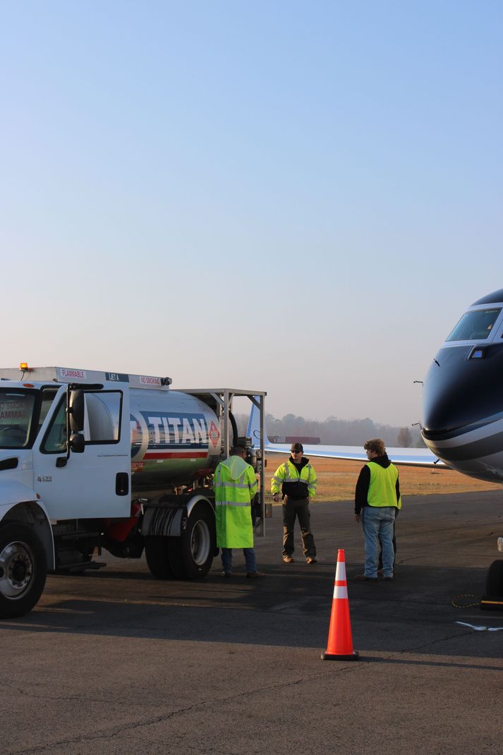 A white Titan fuel truck parked on an airfield next to an airplane, with three personnel in high-visibility vests nearby.