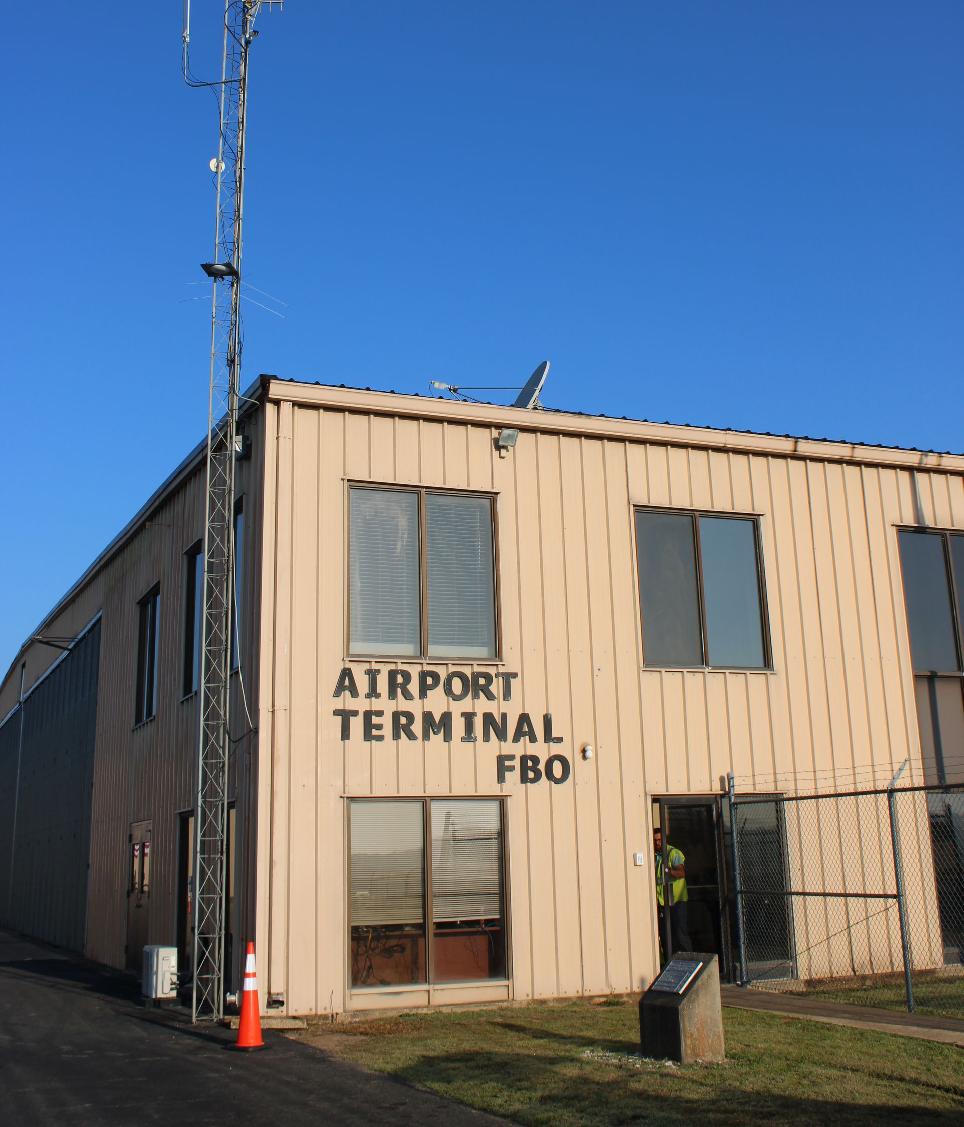 A tan airport terminal building with a communication tower in front, set against a clear blue sky.