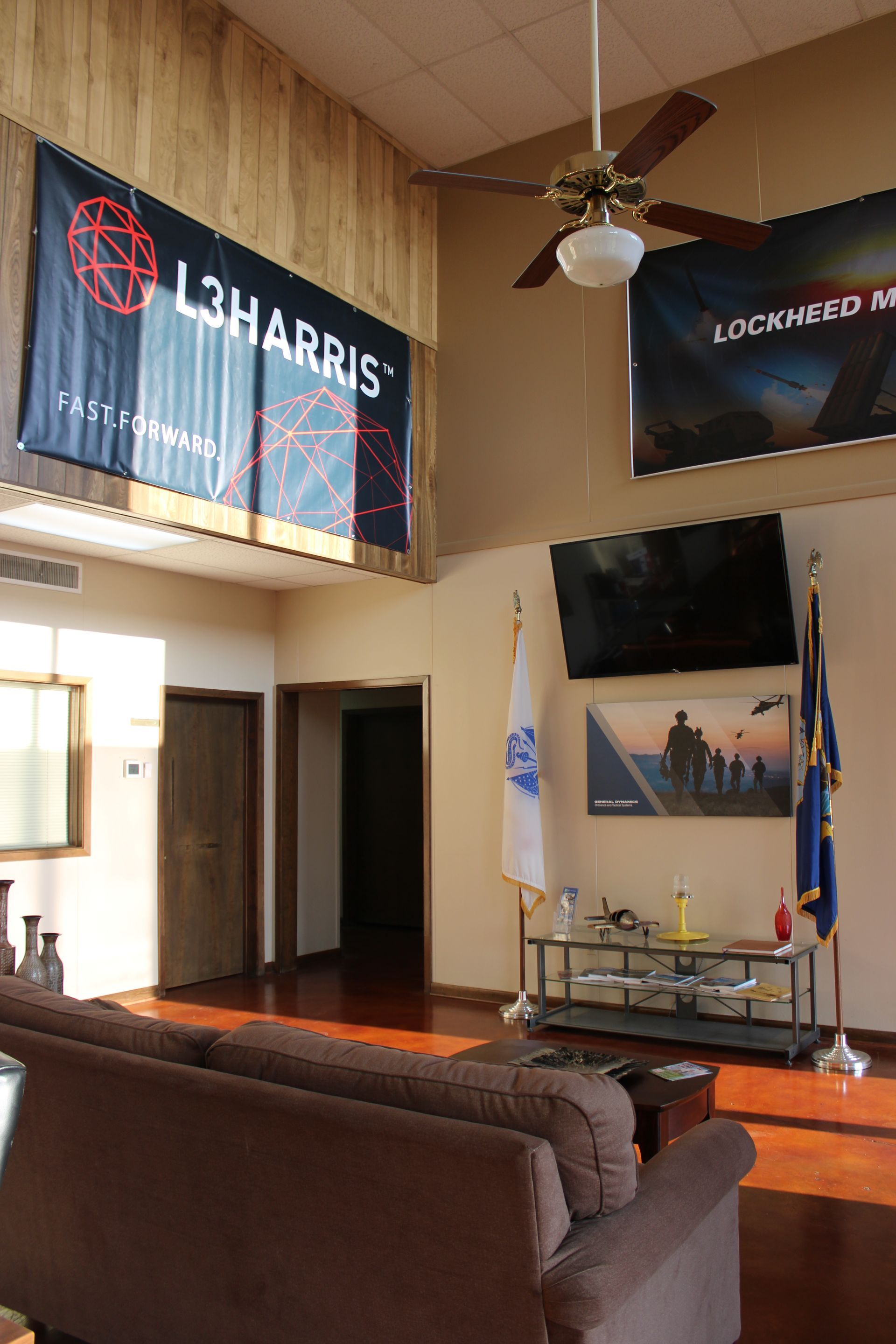 A room with a couch in the foreground, L3Harris and Lockheed Martin banners on walls, flags, and a wall-mounted television.