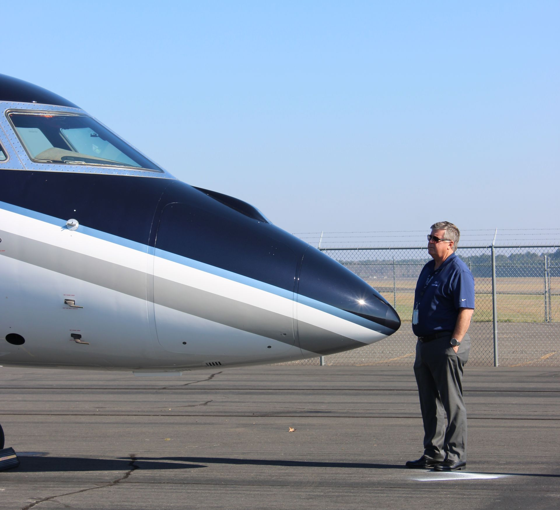 A pilot stands beside a blue-and-white jet on the runway under a clear sky.