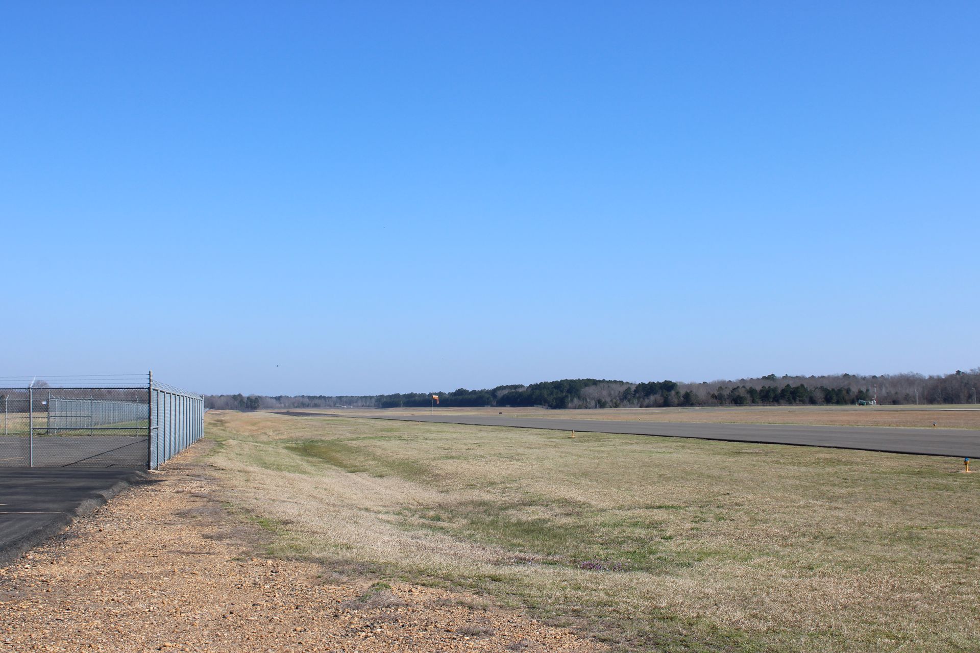 A wide, paved airport runway stretches into the distance under a clear, bright blue sky next to a chain-link fence.