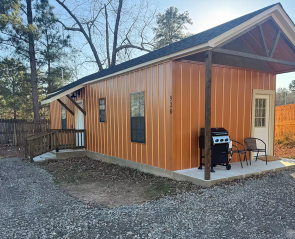 Small orange cabin with a porch, grill, and chair on a gravel lot surrounded by trees