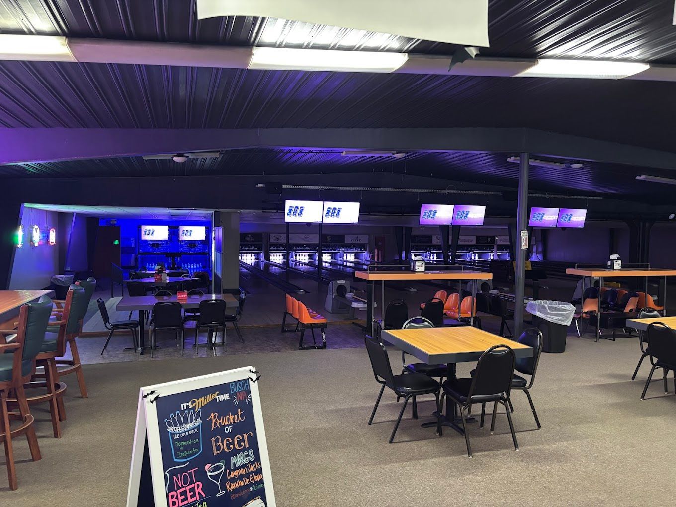 Indoor gaming lounge with tables and chairs, blue-lit arcade area, and overhead TV screens