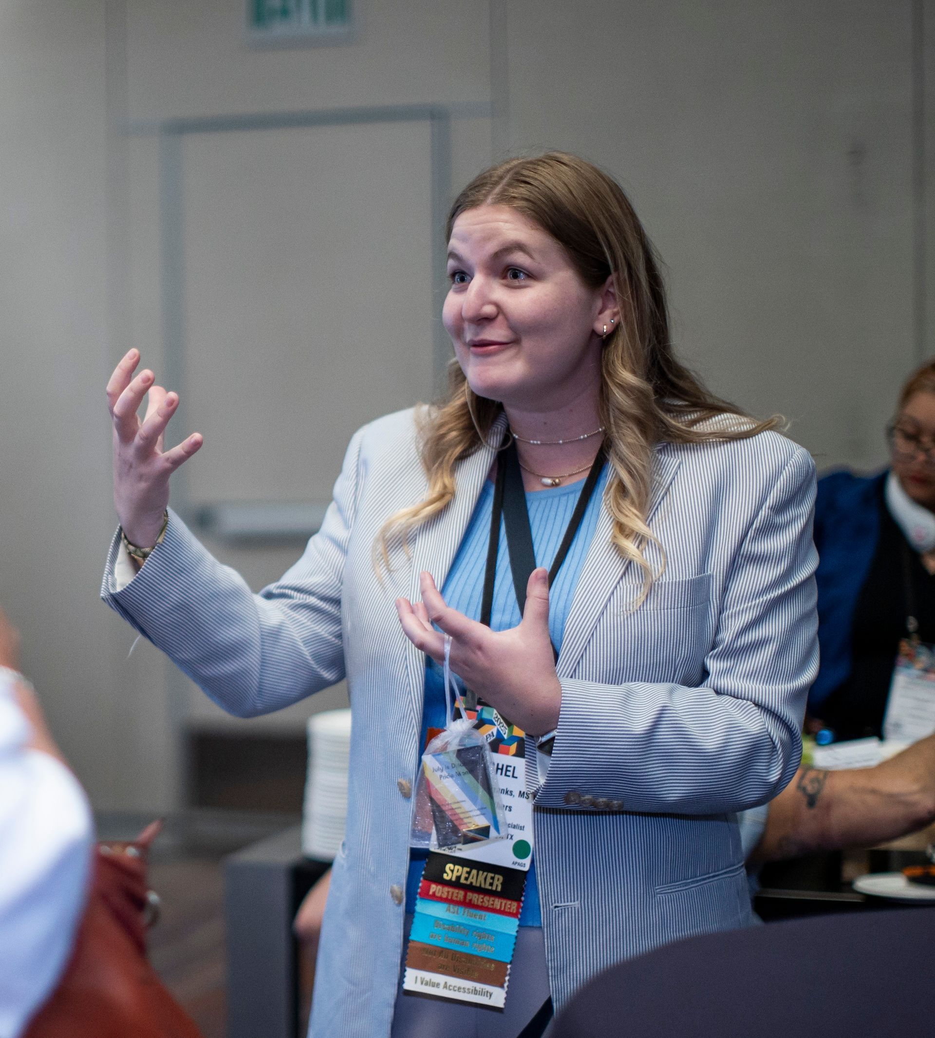 A person using a wheelchair, wearing a colorful patterned top and tan pants, smiling at an event.