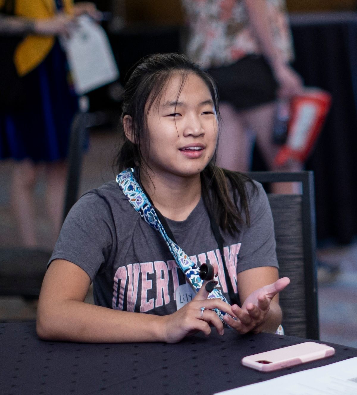 A person wearing a grey shirt and patterned lanyard gestures while speaking at a table with a phone in front of them.