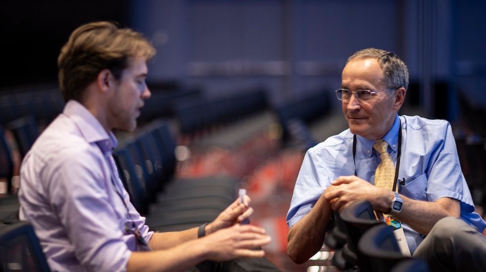 Two men in conversation in a dim auditorium, one gesturing while seated across from the other.