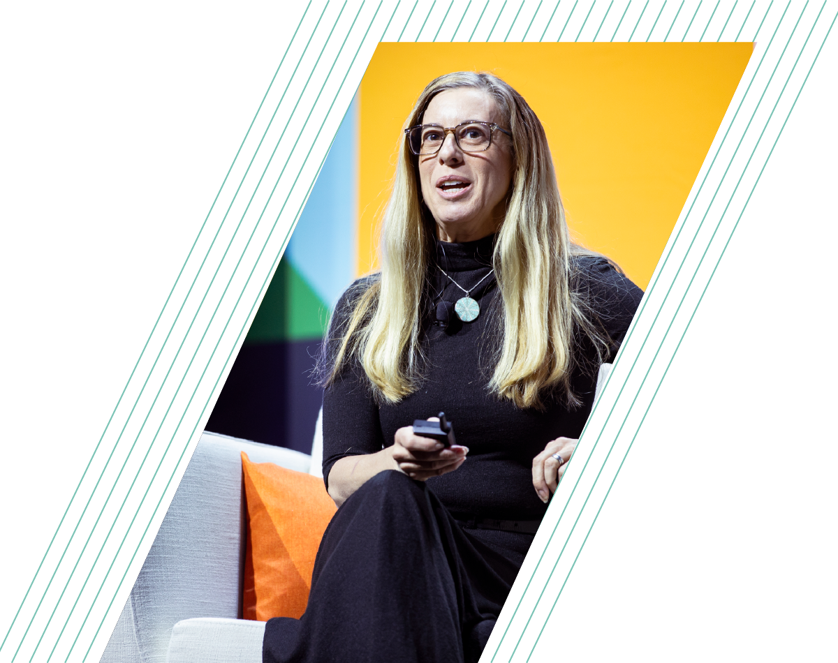 Woman with long blonde hair speaking on a stage, seated on a gray chair against a yellow backdrop.