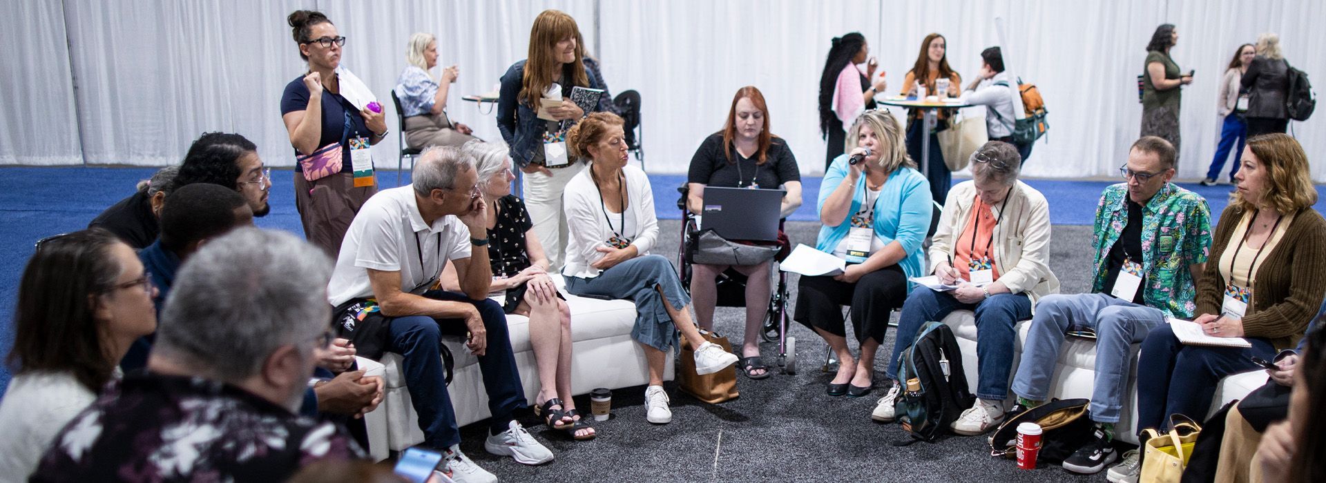 People seated in a circle during a discussion in a bright room, with a facilitator standing nearby.