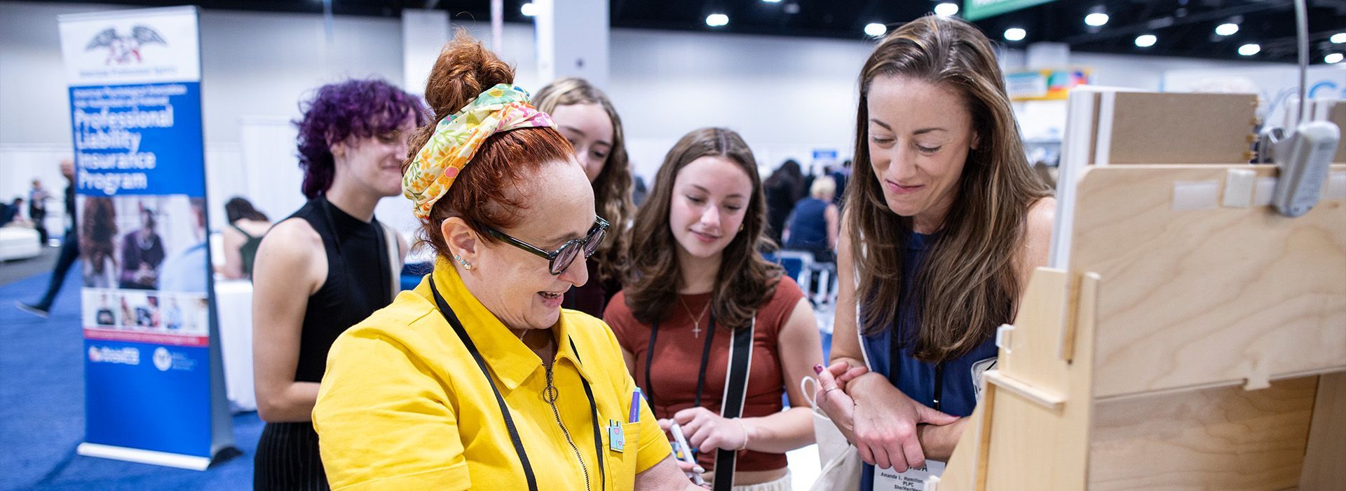 People gathered at a booth, looking at a smartphone with a presenter in a yellow shirt.