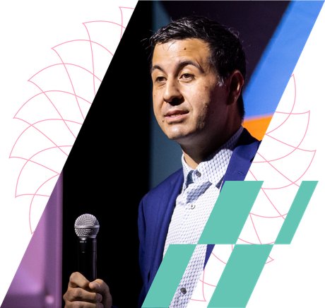 Man speaking into a microphone on a stage, wearing a blue suit with abstract geometric graphics behind him