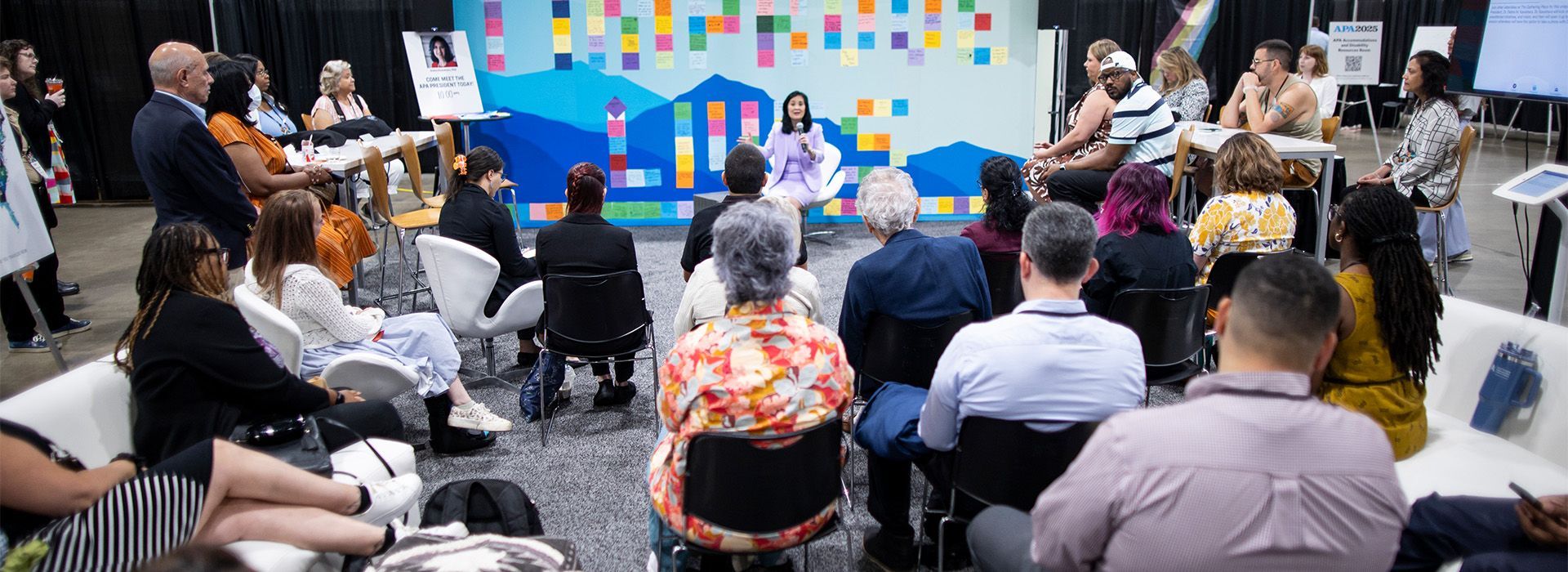 People seated in a meeting room, watching a presentation in front of a blue bulletin board.
