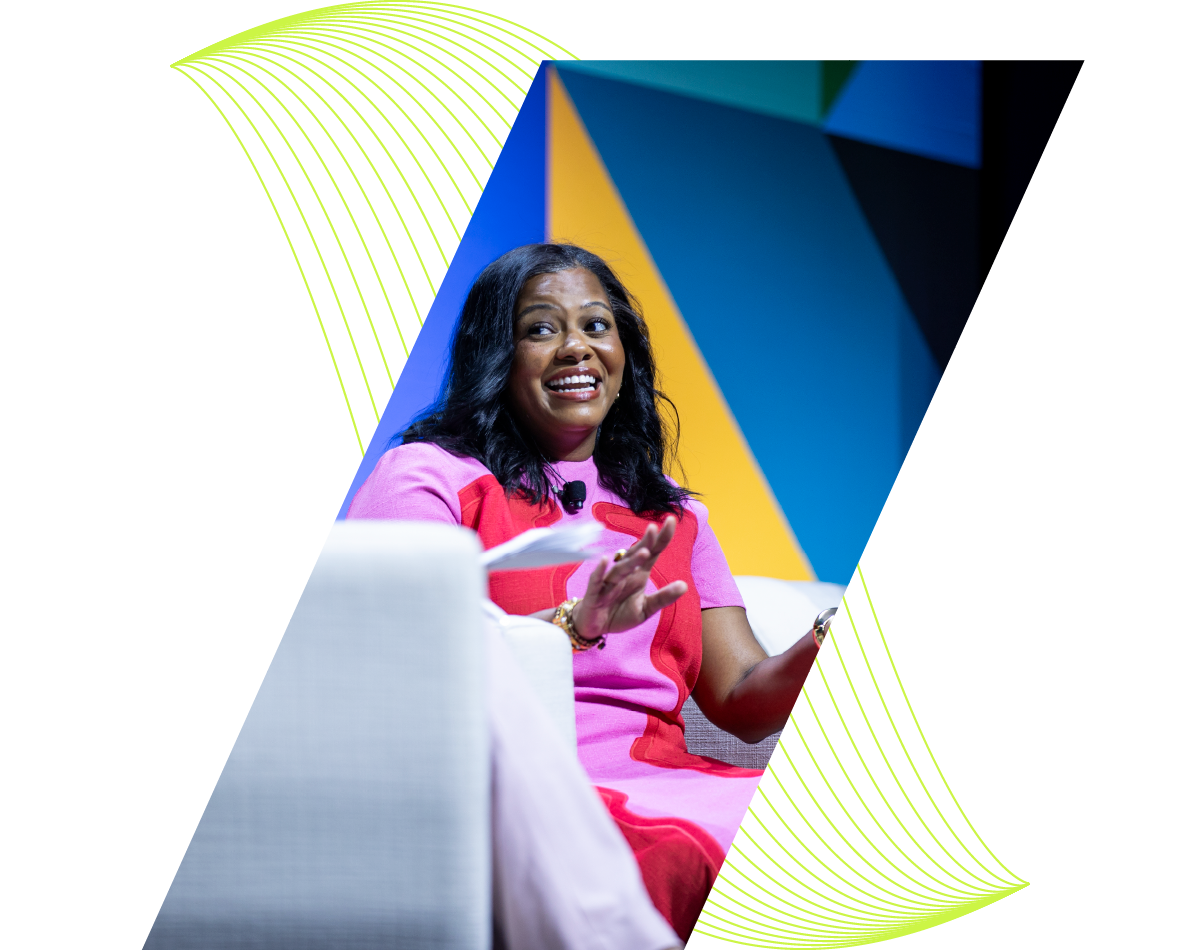 Woman in pink onstage, speaking animatedly against a blue and yellow geometric backdrop.