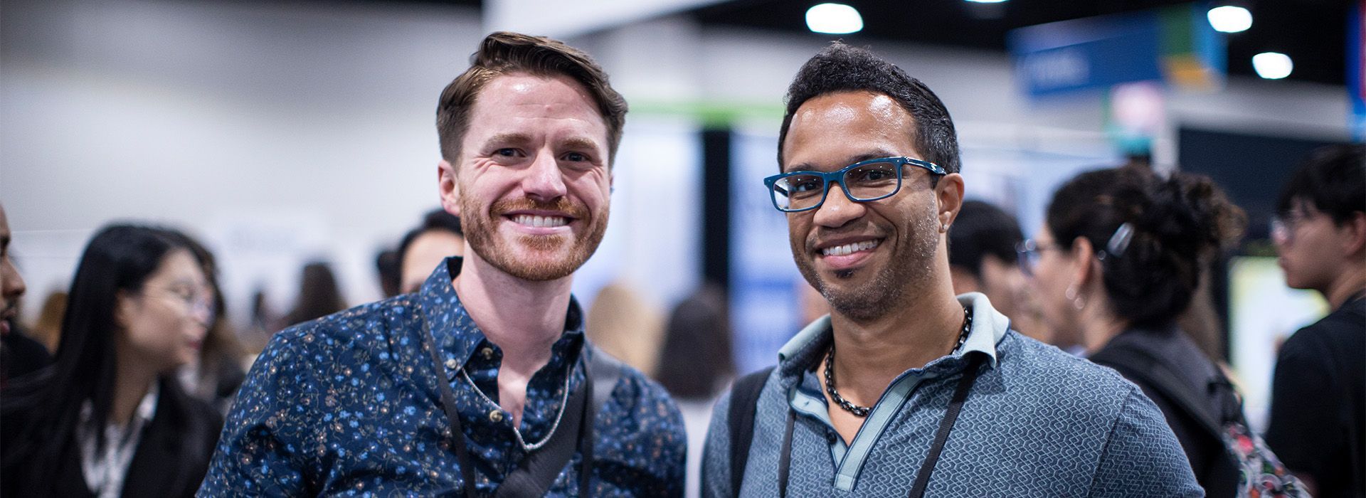 Two smiling men standing together in the solutions center, one in a dark patterned shirt and one in glasses with a lanyard