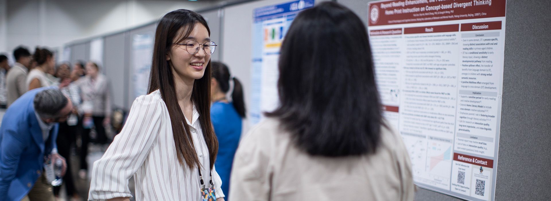 Two women talking in front of a conference poster display, with attendees in the background.