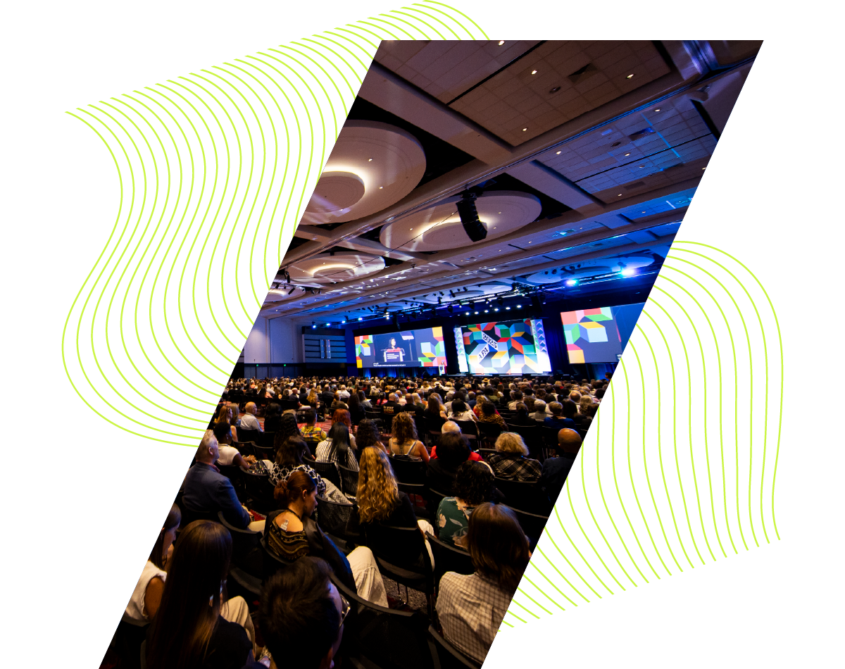 Large conference hall packed with attendees facing a bright stage with multiple screens and blue lighting