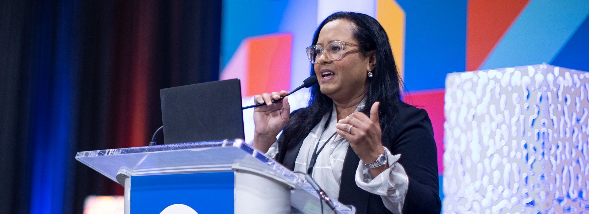 Speaker at podium gesturing during a presentation on a stage with blue and red backdrop
