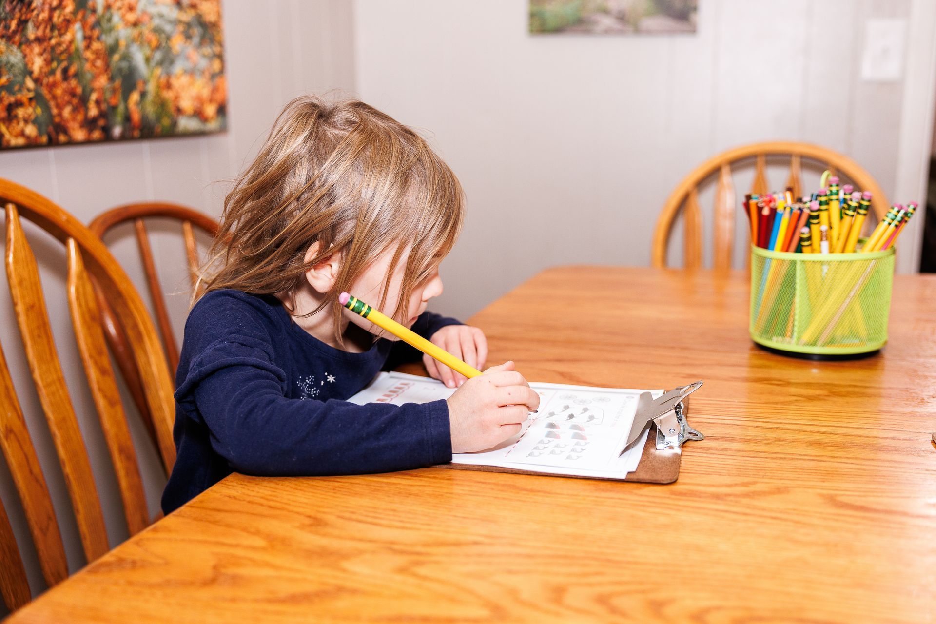 A little girl is sitting at a table with a pencil in her mouth.