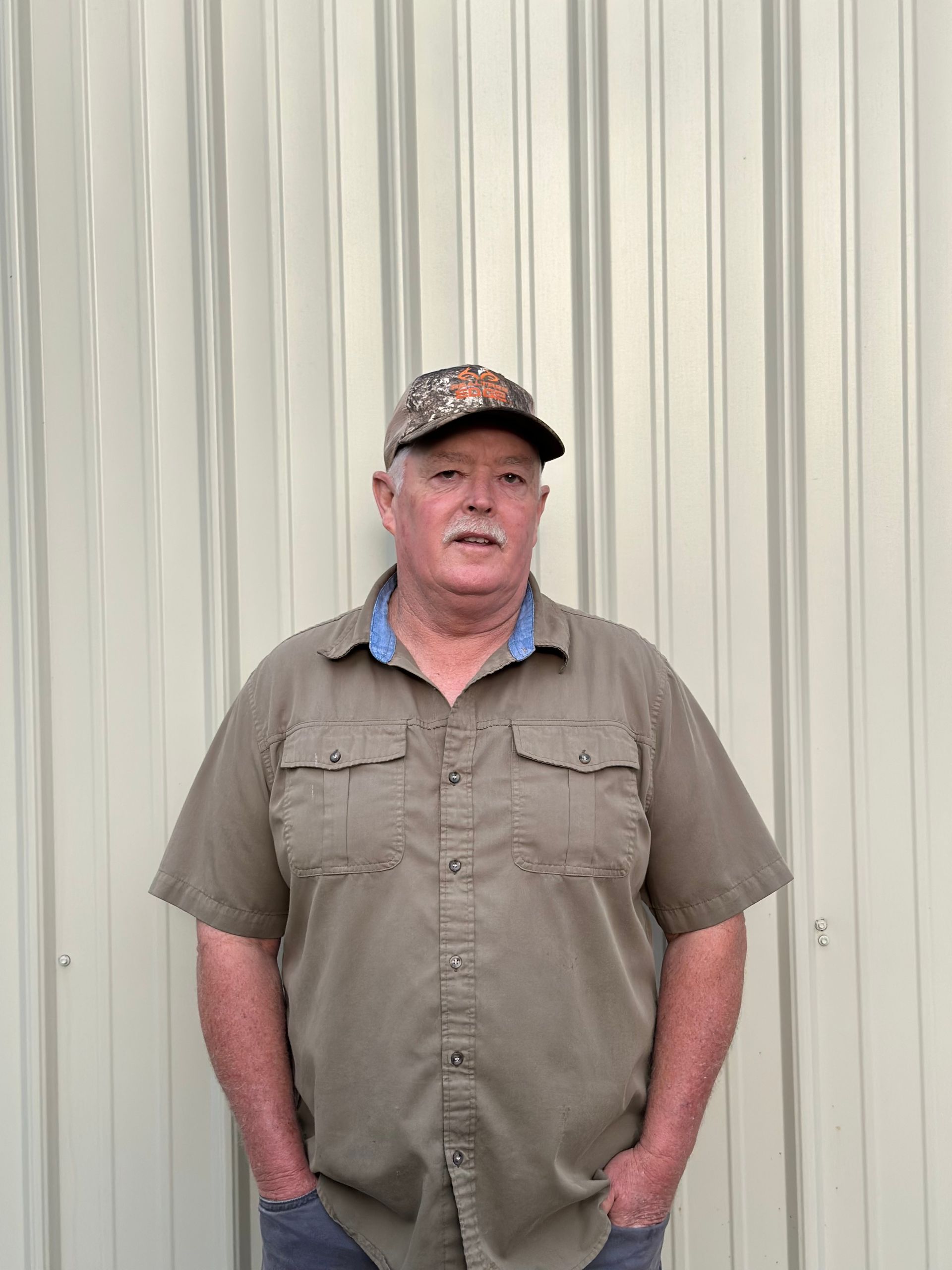 Man wearing a camo hat and tan shirt with hands in pockets, standing in front of a metal wall.