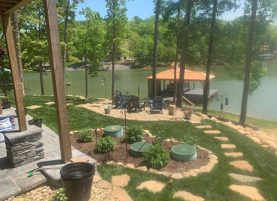 Lakeside yard with stone path and patio, leading to a dock with a gazebo. Trees surround the water.