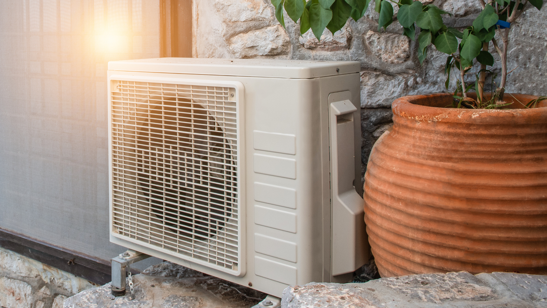 A white air conditioner is sitting on the side of a building next to a potted plant.