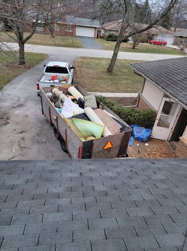 Dump trailer filled with debris parked in a driveway beside a house, with a white truck nearby.