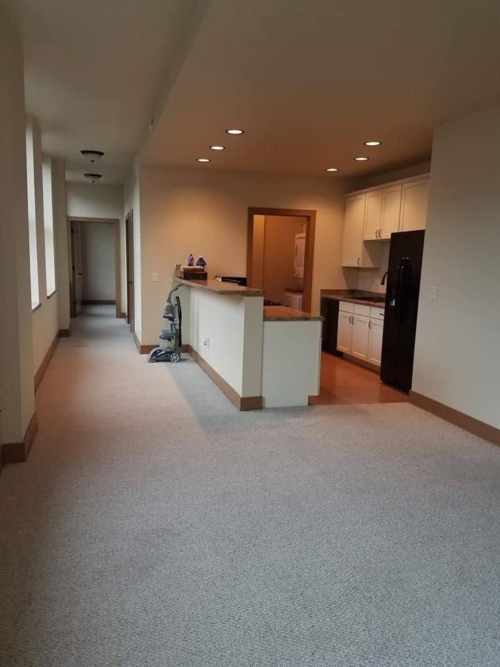 Empty apartment hallway opening to a kitchen with white cabinets and a black refrigerator.