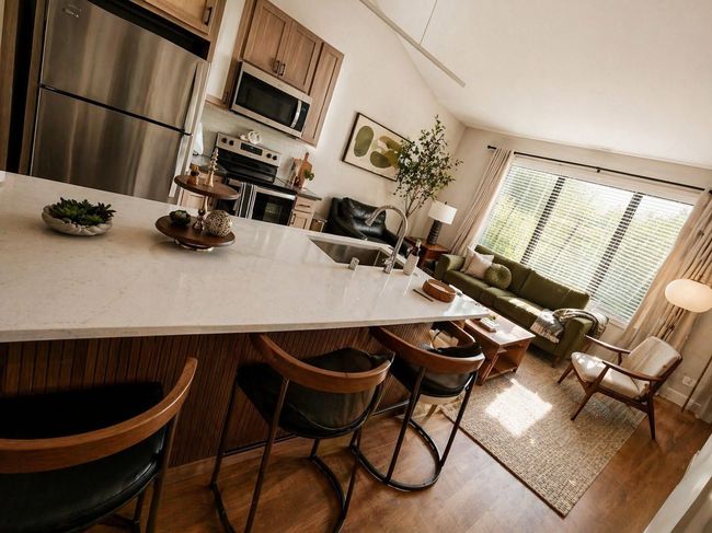 Modern kitchen with white island, bar stools, stainless fridge, and sunlit living area beyond.