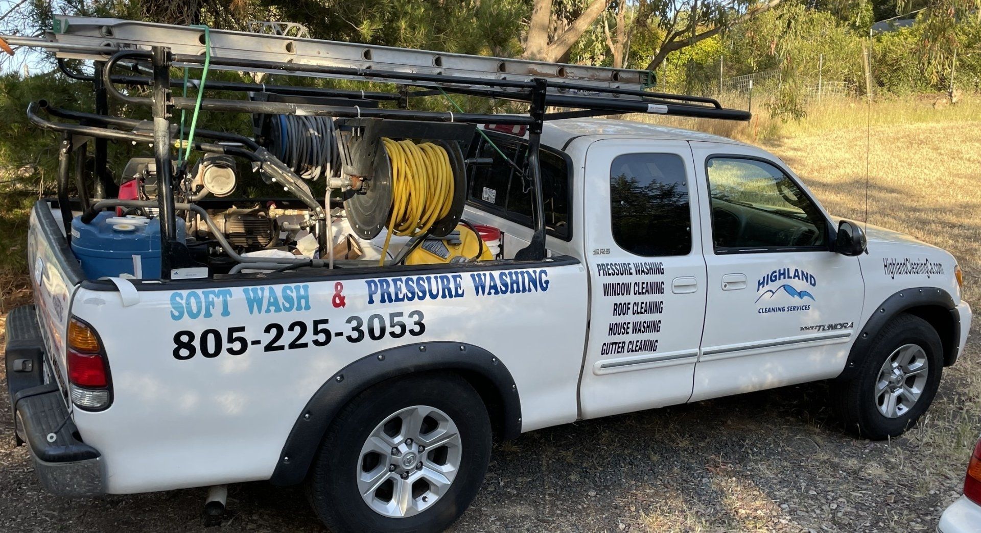 A white truck with a ladder on the back is parked in a gravel lot.
