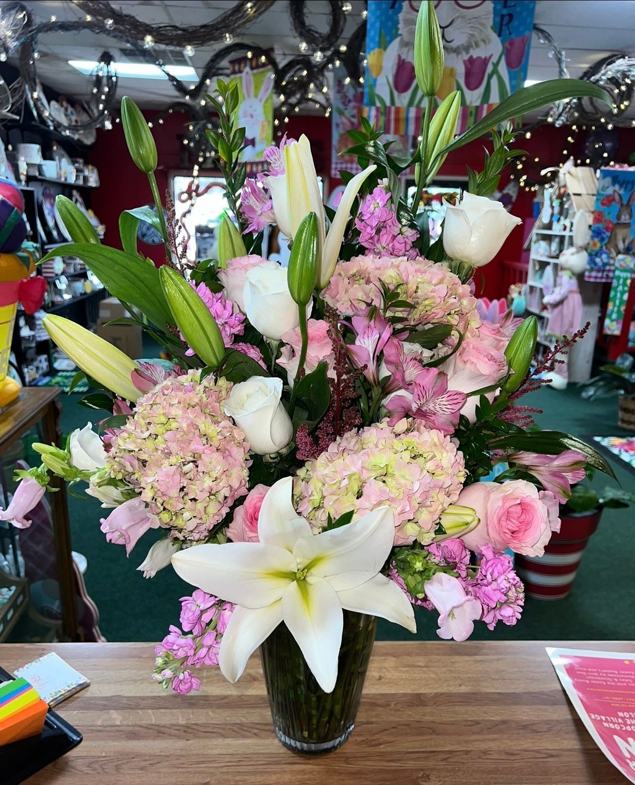 Pink and white floral arrangement in a clear vase on a wood surface; hydrangeas, lilies, roses.