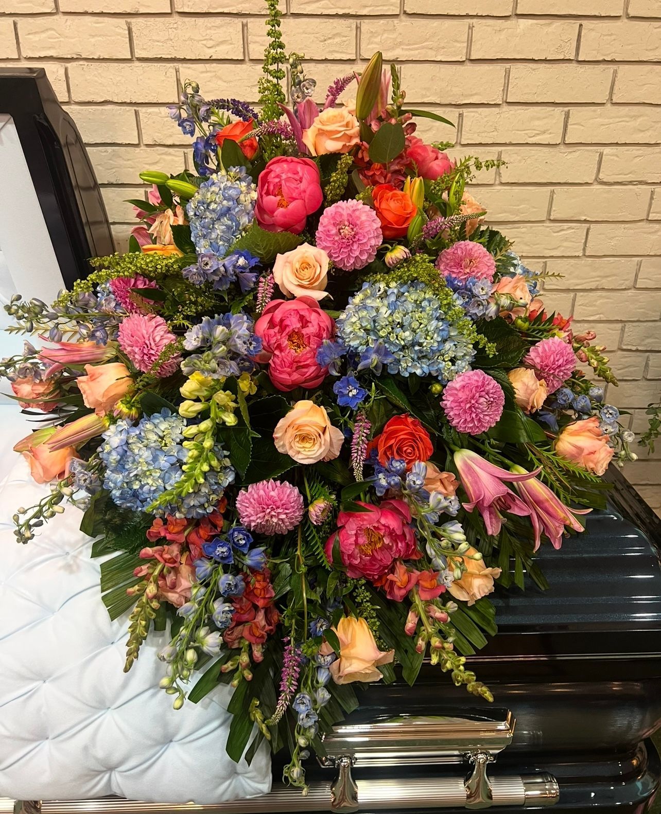 Floral arrangement of bright pink, blue, and peach flowers on a dark casket, with a brick wall backdrop.