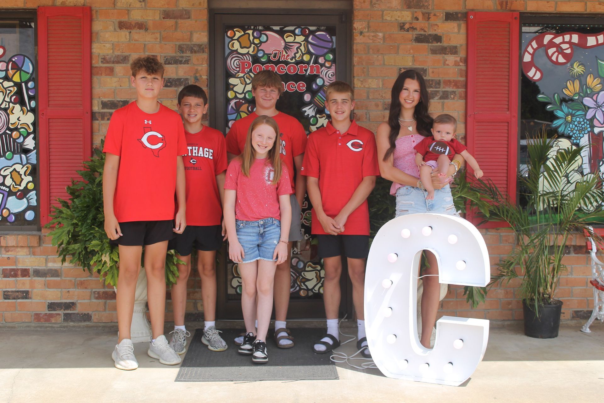 Group of people in front of a store, holding a large "C." Many wear red shirts and some hold a baby.