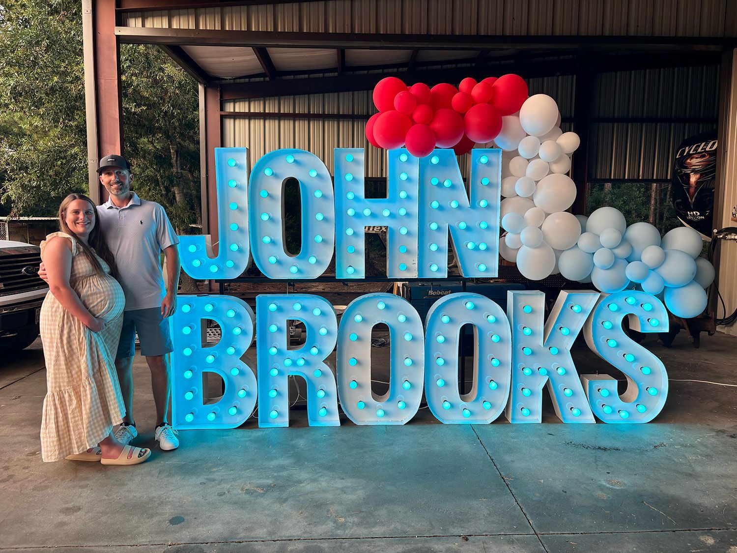 Pregnant couple poses next to illuminated "JOHN BROOKS" letters, with red, white, and blue balloon arch.