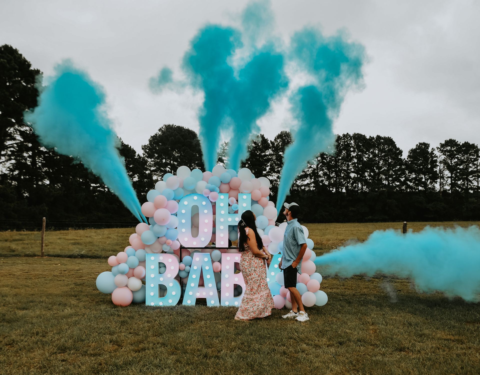 Couple at gender reveal, blue smoke erupts from balloons spelling "OH BABY" in field.