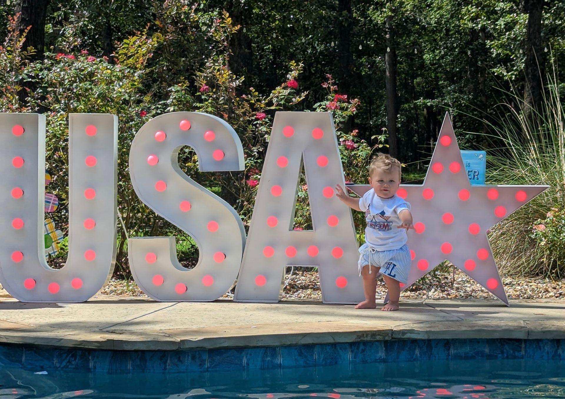 Child stands near a pool with illuminated "USA" letters and a star. Bright sunny day.