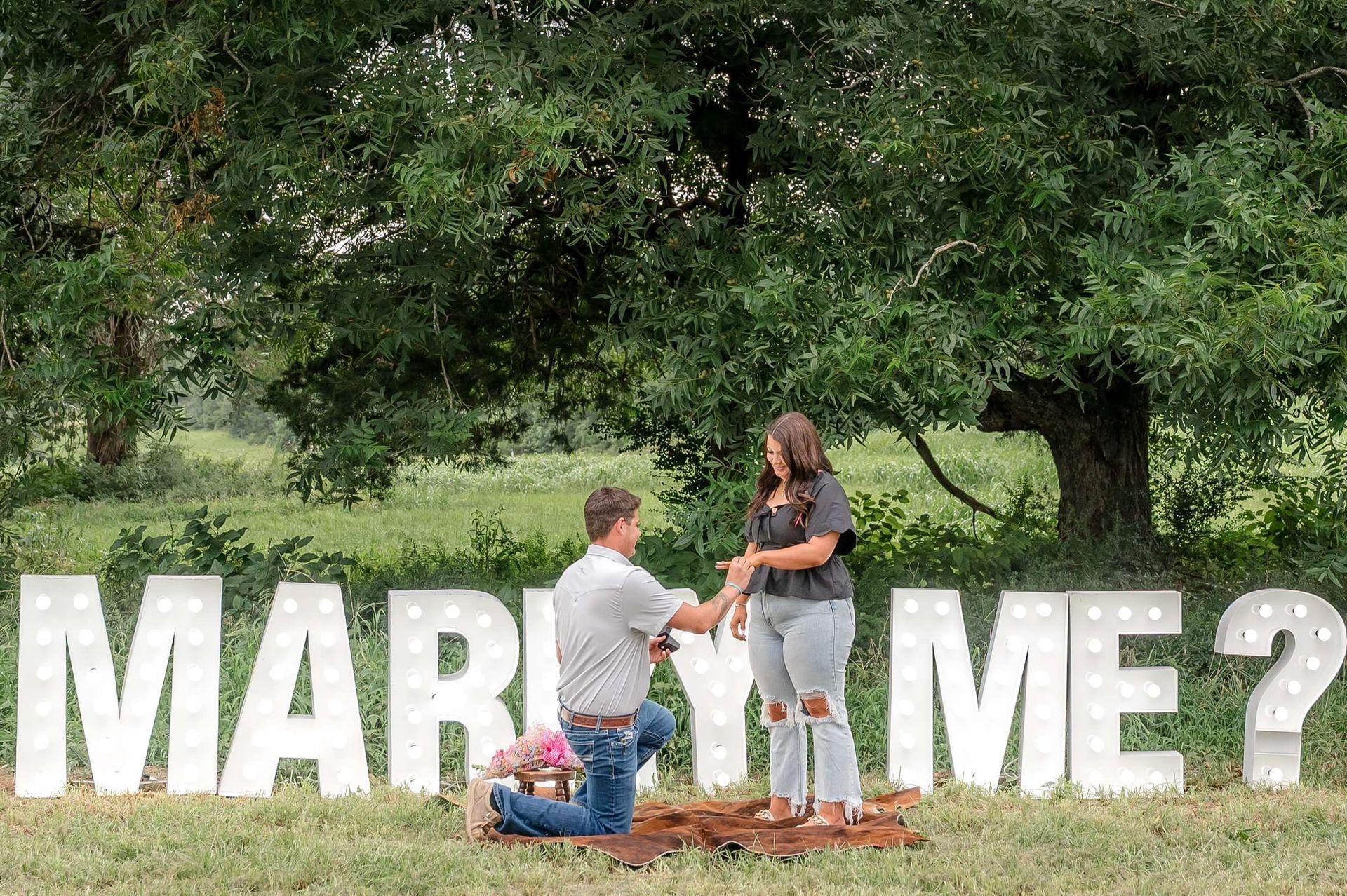 Man on one knee proposing to woman with "MARRY ME?" letters in a field.