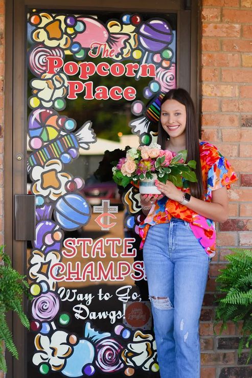 Woman with flowers outside Popcorn Place door, decorated with candy. 