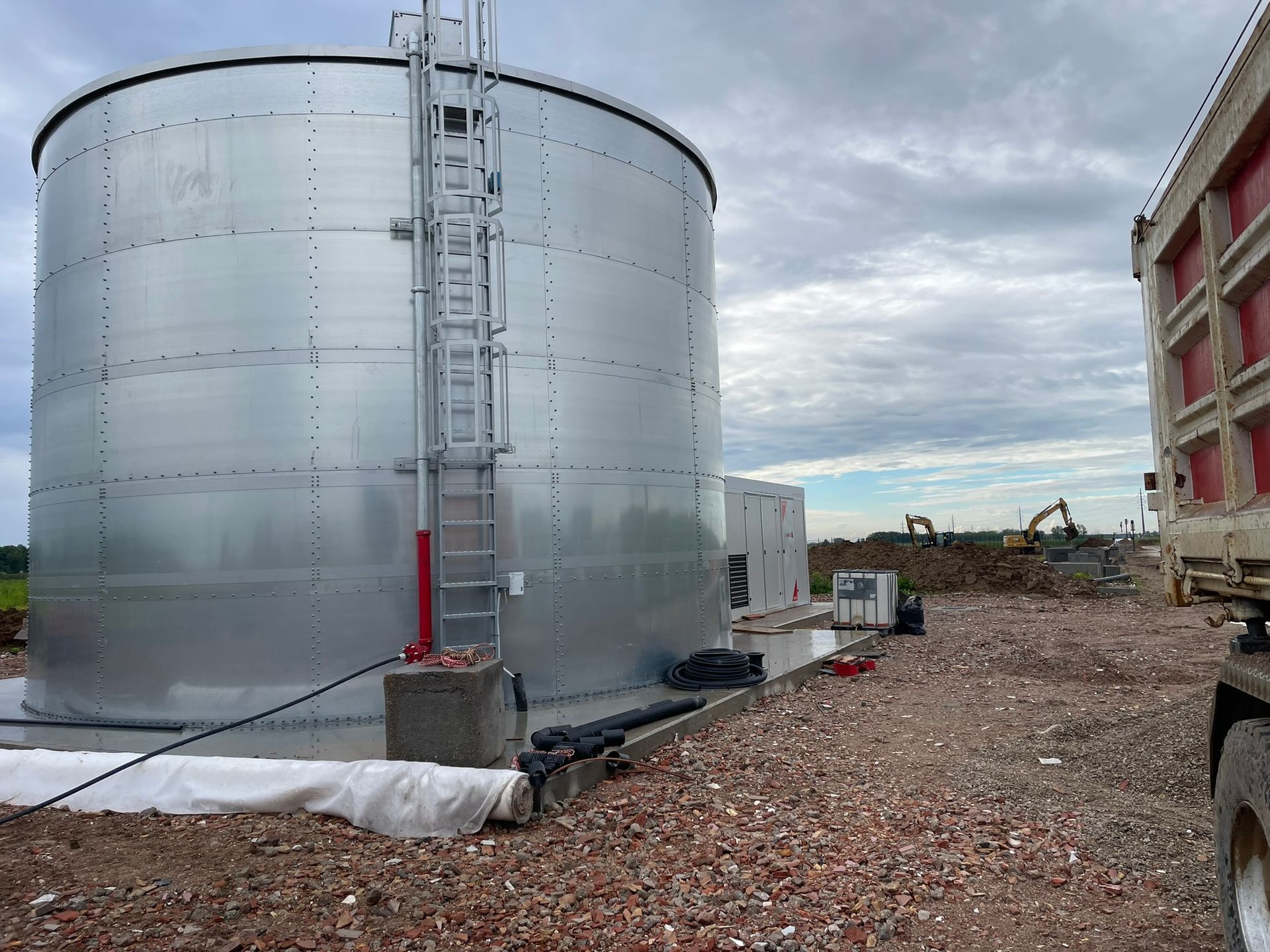 Un grande serbatoio di metallo è appoggiato su un terreno ghiaioso accanto a un autocarro con cassone ribaltabile.