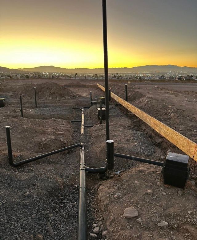 Utility pipes and conduit at a dirt construction site at sunset, with mountains in the distance.