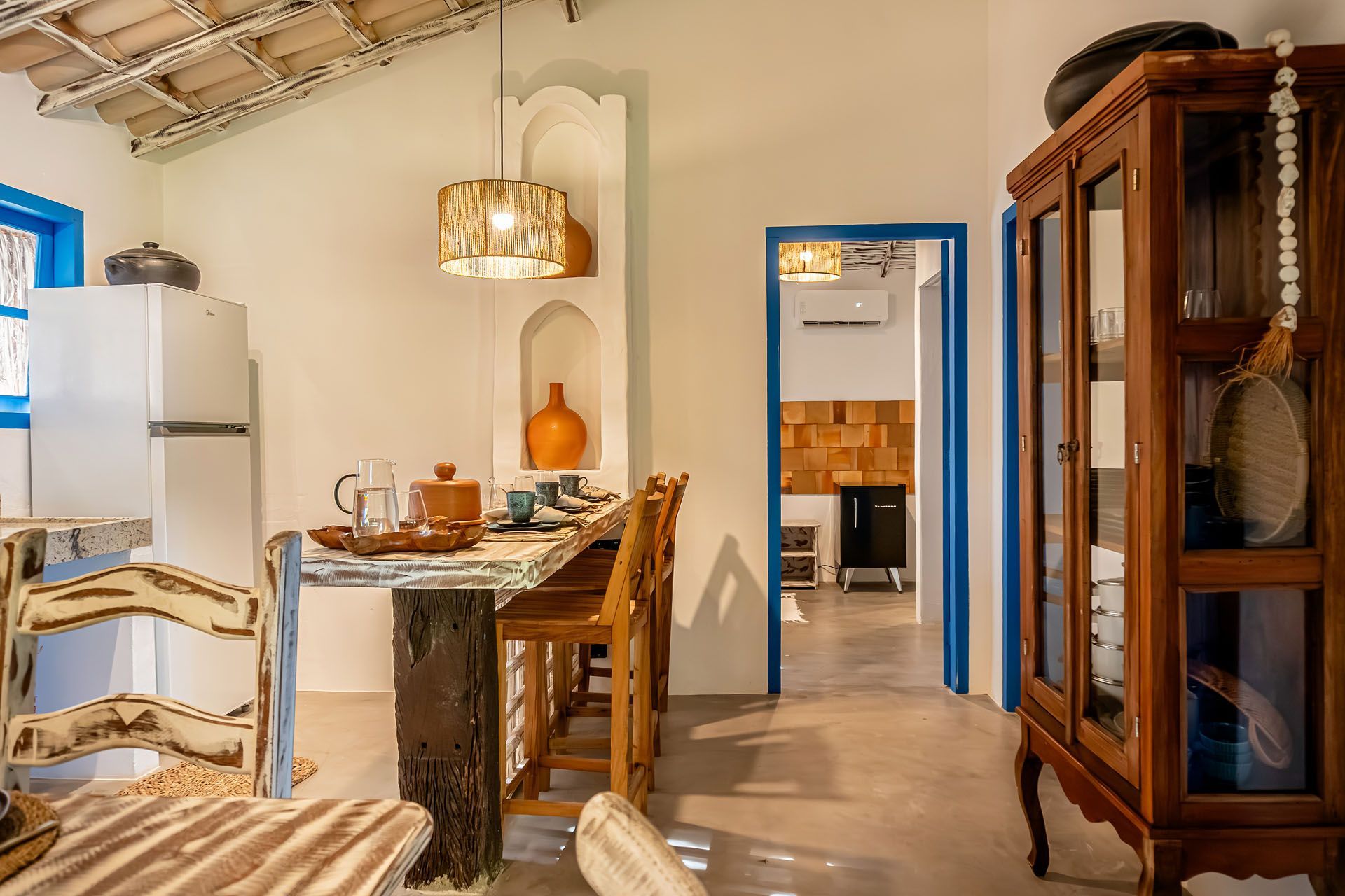 Sala de jantar rústica e aconchegante com mesa de madeira, cadeiras de vime, luminária pendente e porta com detalhes em azul.