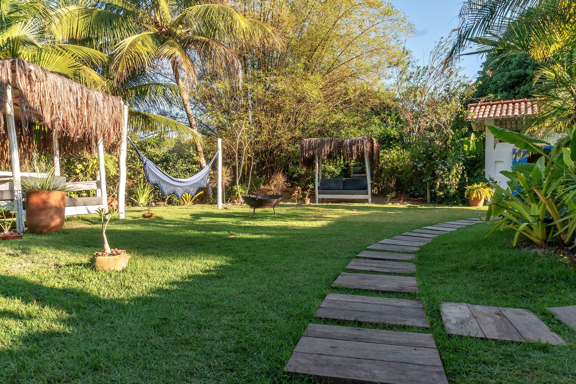 Jardim ensolarado com gramado, rede, árvores e caminho de pedras que leva a um pequeno pátio.