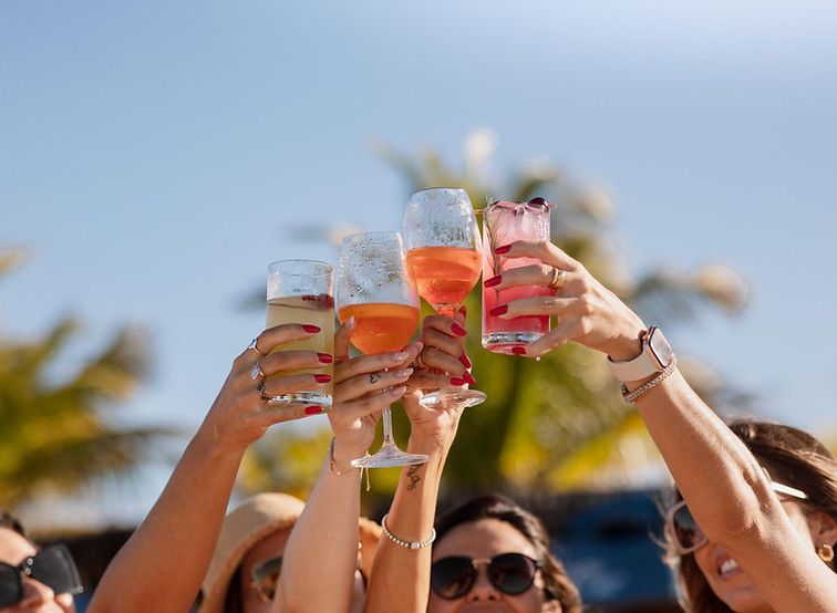 Amigos brindam com bebidas coloridas em um evento ensolarado ao ar livre, à beira da piscina.