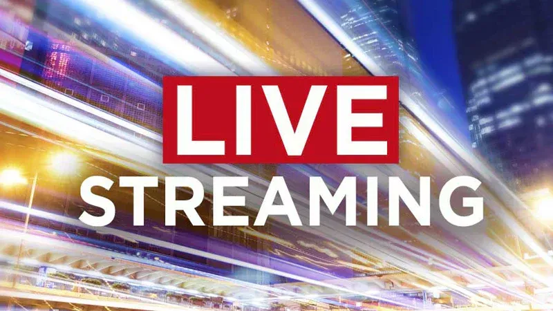 Two people filming an event with cameras on tripods. One sits, the other stands. Blue background, blurry crowd.