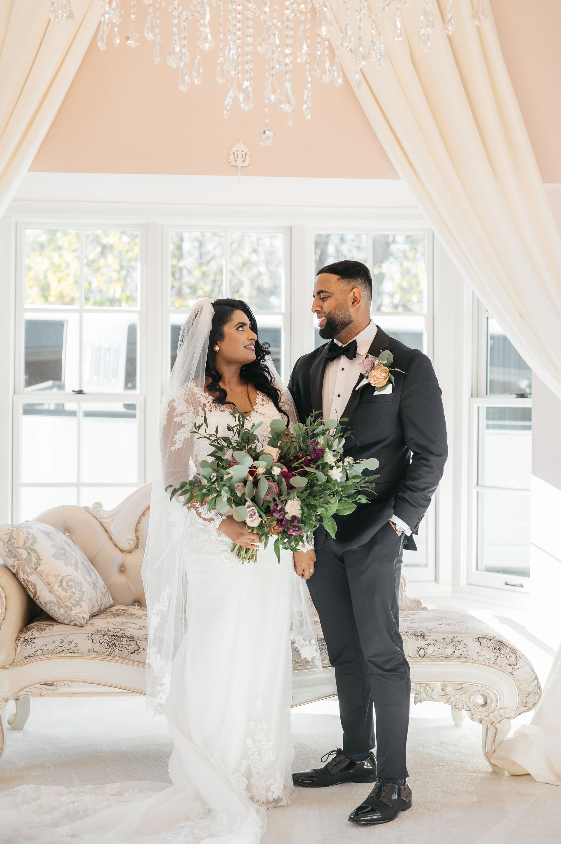 Bride and groom embrace in a field, holding flowers. She wears a white gown and veil; he wears a blue suit.