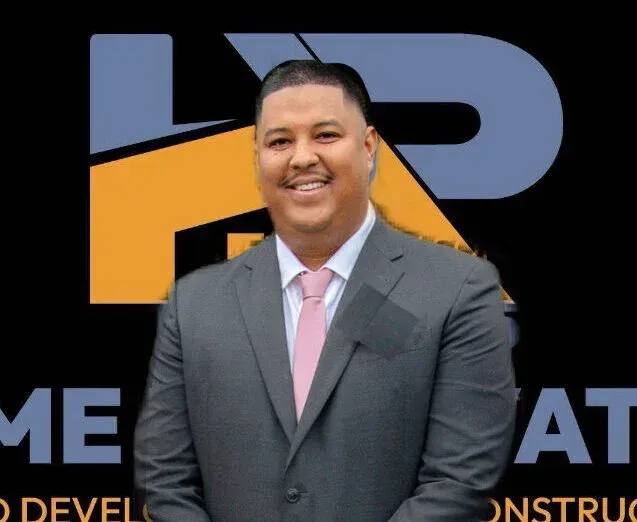 Man in a suit smiles, in front of a company logo with yellow and blue colors.