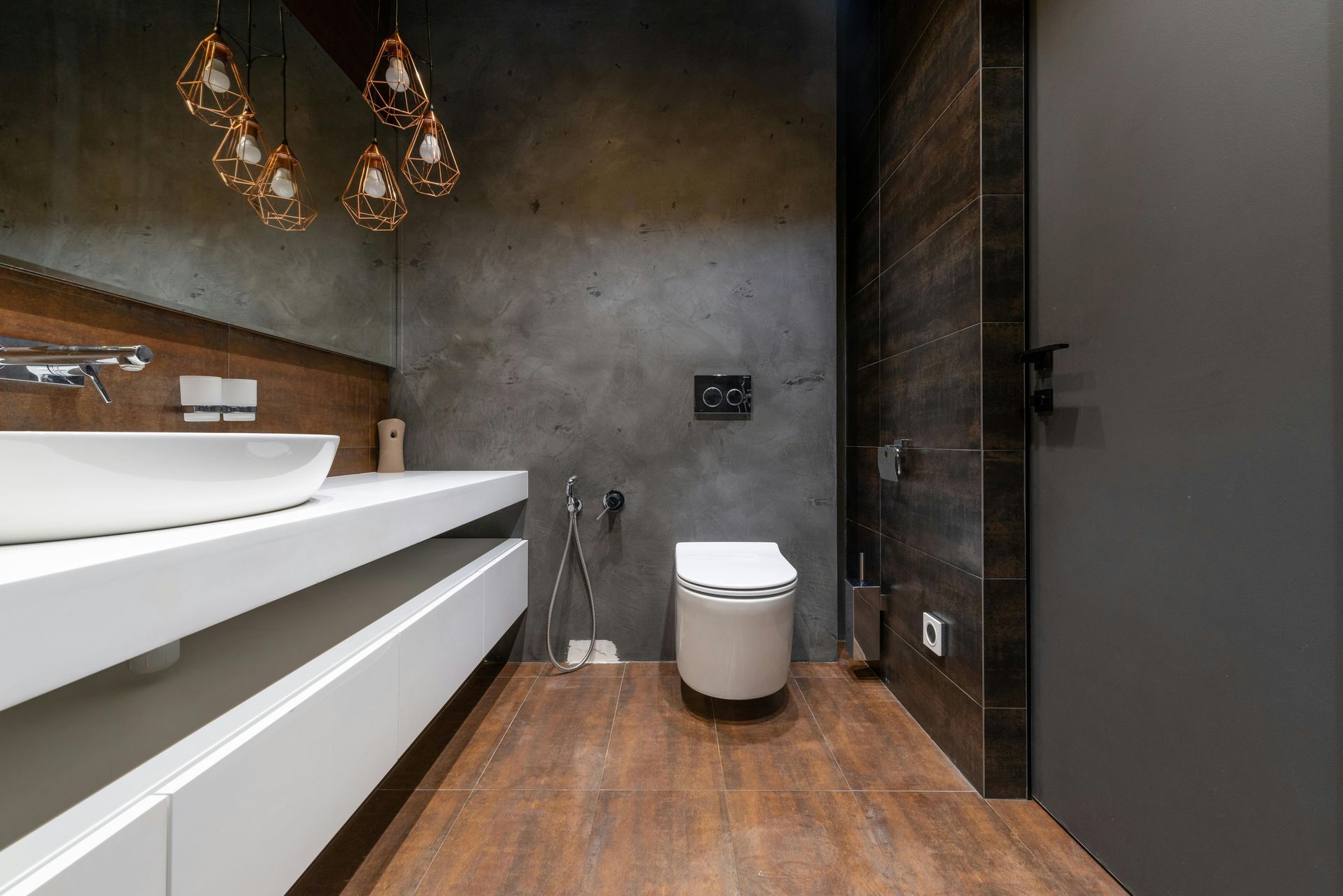 Modern bathroom with floating sink, toilet, dark walls, wood floor, and decorative lights.