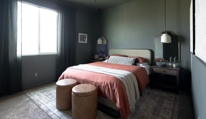 A bedroom with a bed , two ottomans , a mirror and a window.