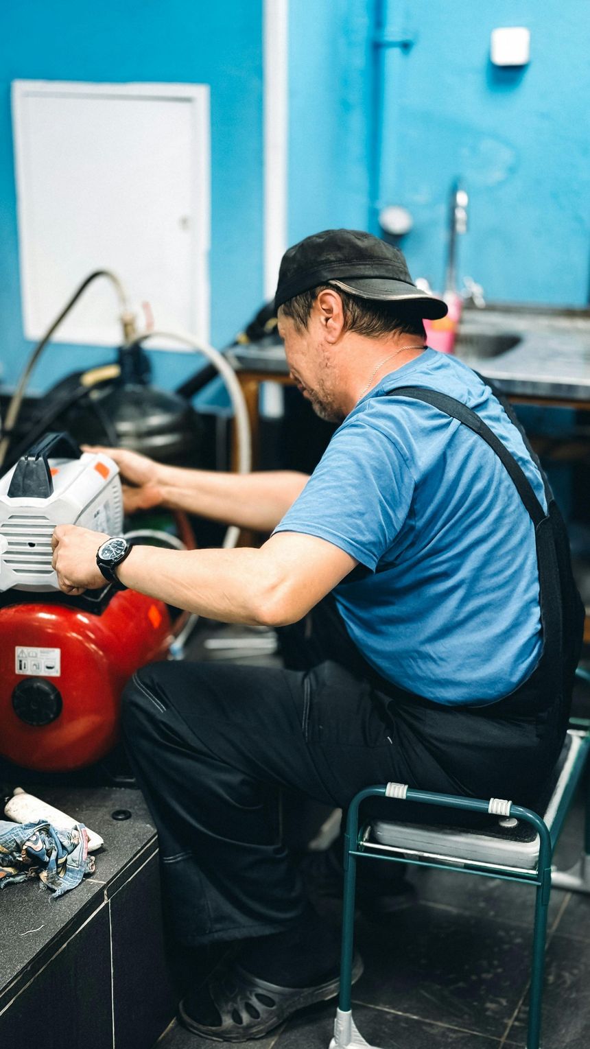 Man in blue shirt working on machinery; red tank, dark cap, metal shop setting.