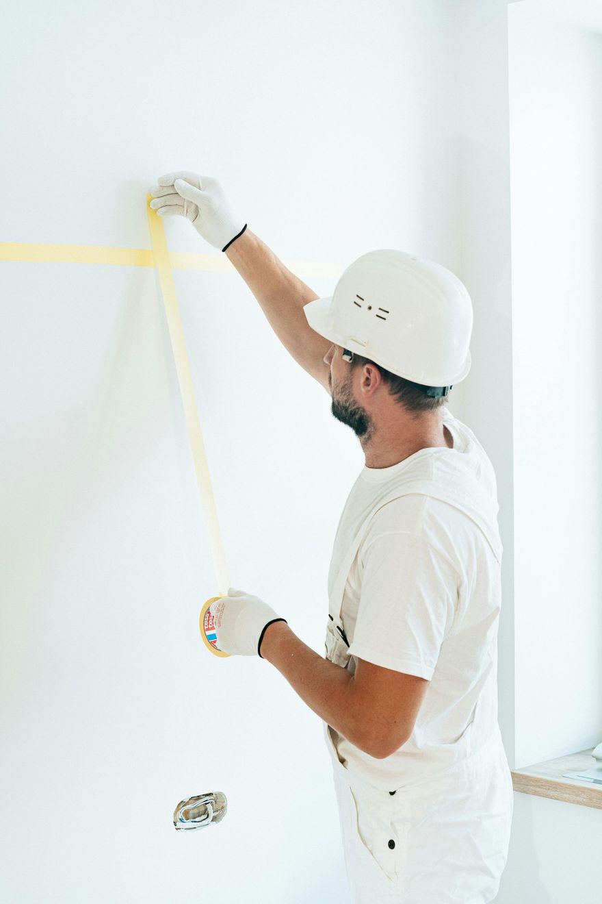 Person in white overalls and hard hat applies painter's tape to a wall.